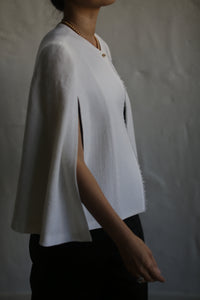 A person wearing the Capelet Long in Japanese Wool Gauze Undyed over a black outfit is shown in a side profile. The background features a neutral, slightly textured wall, and the lighting accentuates the drape and texture of the fabric.