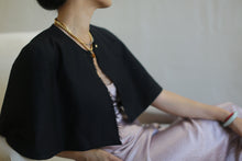 Load image into Gallery viewer, A person with short dark hair sits on a white chair, wearing a black Japanese Wool Gauze Capelet over a light pink satin dress, and a gold necklace. The custom item was crafted for this specific date. The person&#39;s face is turned away from the camera, and their right hand rests on their lap. The background is neutral.