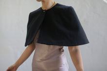 Load image into Gallery viewer, A person is wearing a Capelet | Japanese Wool Gauze Black over a light-colored dress. The image is cropped at the shoulders, leaving their face not visible. They are posed against a plain, light grayish background. A gold necklace, which appears to be custom-made, is visible around their neck.