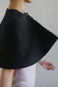 Close-up of an individual wearing the Capelet | Japanese Wool Gauze Black, with the garment flowing elegantly to the side. The person’s arm, adorned with bracelets, peeks from beneath a light-colored shirt. The black capelet, handcrafted by Atelier Lan Jaenicke from luxurious Japanese wool gauze, contrasts beautifully against the soft neutral background.