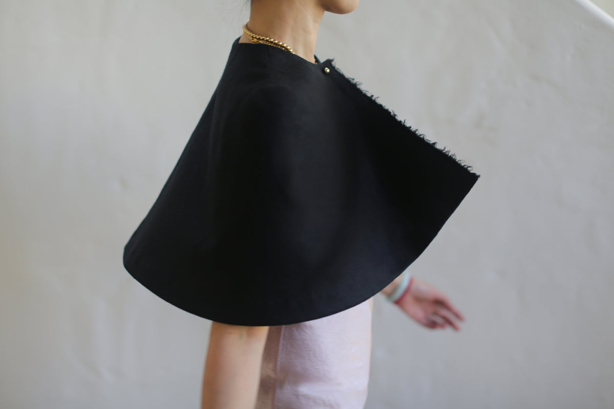 Profile view of a person wearing the "Capelet | Japanese Wool Gauze Black" with fringed edges over a light-colored outfit, set against a plain, light-colored background. The made-to-order capelet is adorned with a gold necklace, showcasing impeccable craftsmanship and style.