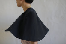 Load image into Gallery viewer, A person with a bun hairstyle is wearing the Capelet | Japanese Wool Gauze Black, paired with a gold necklace. They are captured in a side profile against a neutral, blurred background, giving the image a sense of motion. This unique ensemble appears to be custom-made to order, showcasing personalized fashion at its finest.