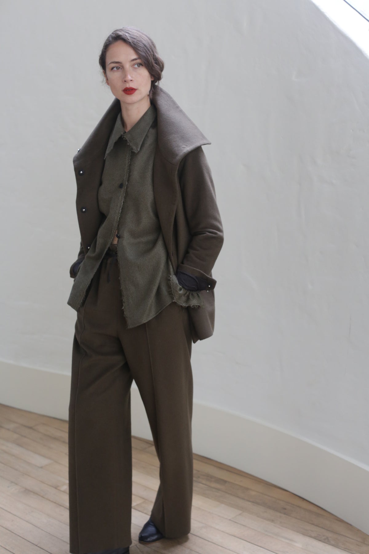 A person stands indoors against a light-colored wall, wearing an olive-green outfit with a coat, buttoned shirt, and Cashmere Trousers | Olive. They have dark hair pulled to one side and red lipstick. The setting includes a wooden floor and white baseboards, subtly complemented by silk ribbon ties on the coat.