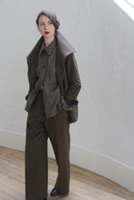 Load image into Gallery viewer, A person stands indoors against a light-colored wall, wearing an olive-green outfit with a coat, buttoned shirt, and Cashmere Trousers | Olive. They have dark hair pulled to one side and red lipstick. The setting includes a wooden floor and white baseboards, subtly complemented by silk ribbon ties on the coat.