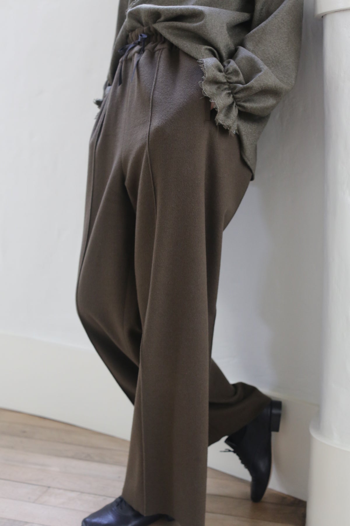 Person leaning against a wall, wearing loose-fitting Olive Cashmere Trousers and a greenish-gray fringed top with silk ribbon ties. Their feet are crossed and they are wearing black shoes. The photo is cropped to show only the torso and legs, emphasizing the relaxed and casual posture.
