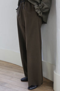 The image shows the lower half of a person standing indoors. They are wearing Cashmere Trousers | Olive with silk ribbon ties, a frayed-hem sweater, and black shoes. The background consists of a white painted wall and light-colored wooden floor.