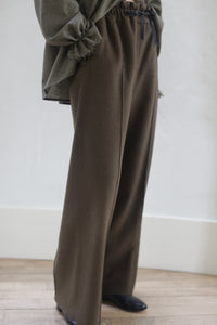 The image shows a person standing in Cashmere Trousers | Olive, paired with a loose, long-sleeved olive green top. The floor is wooden and the background features a white wall. The person is partially cropped out of the frame.