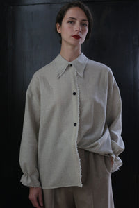 A woman stands against a dark background, wearing a loose-fitting, beige Painter's Shirt - Japanese Wool Gauze | Natural and matching beige pants made from Japanese wool gauze. She has short hair and wears a serious expression with red lipstick. One hand is in her pocket, and her shirt has a frayed edge along the buttons, showcasing sustainable design.