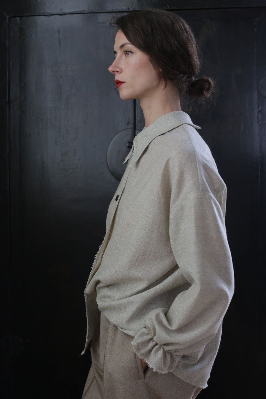 A woman with dark hair tied in a low bun stands against a black background, facing to the left. She wears a light beige oversized Painter's Shirt - Japanese Wool Gauze | Natural and matching pants made from Japanese wool gauze. Her expression is calm and composed, reflecting the sustainable design of her handmade attire.