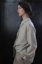 Load image into Gallery viewer, A woman with dark hair tied in a low bun stands against a black background, facing to the left. She wears a light beige oversized Painter&#39;s Shirt - Japanese Wool Gauze | Natural and matching pants made from Japanese wool gauze. Her expression is calm and composed, reflecting the sustainable design of her handmade attire.