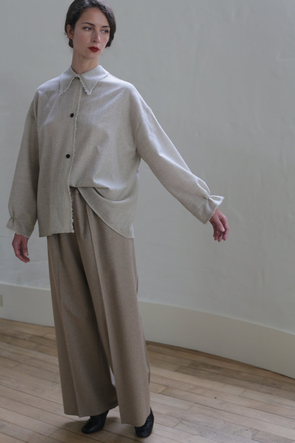 A person with short dark hair is posing inside a minimalistic room. They are wearing the Painter's Shirt - Japanese Wool Gauze | Natural with wide sleeves and light brown wide-leg pants. The expression is neutral, and one arm is slightly extended outward. The floor is wooden, emphasizing the sustainable design.