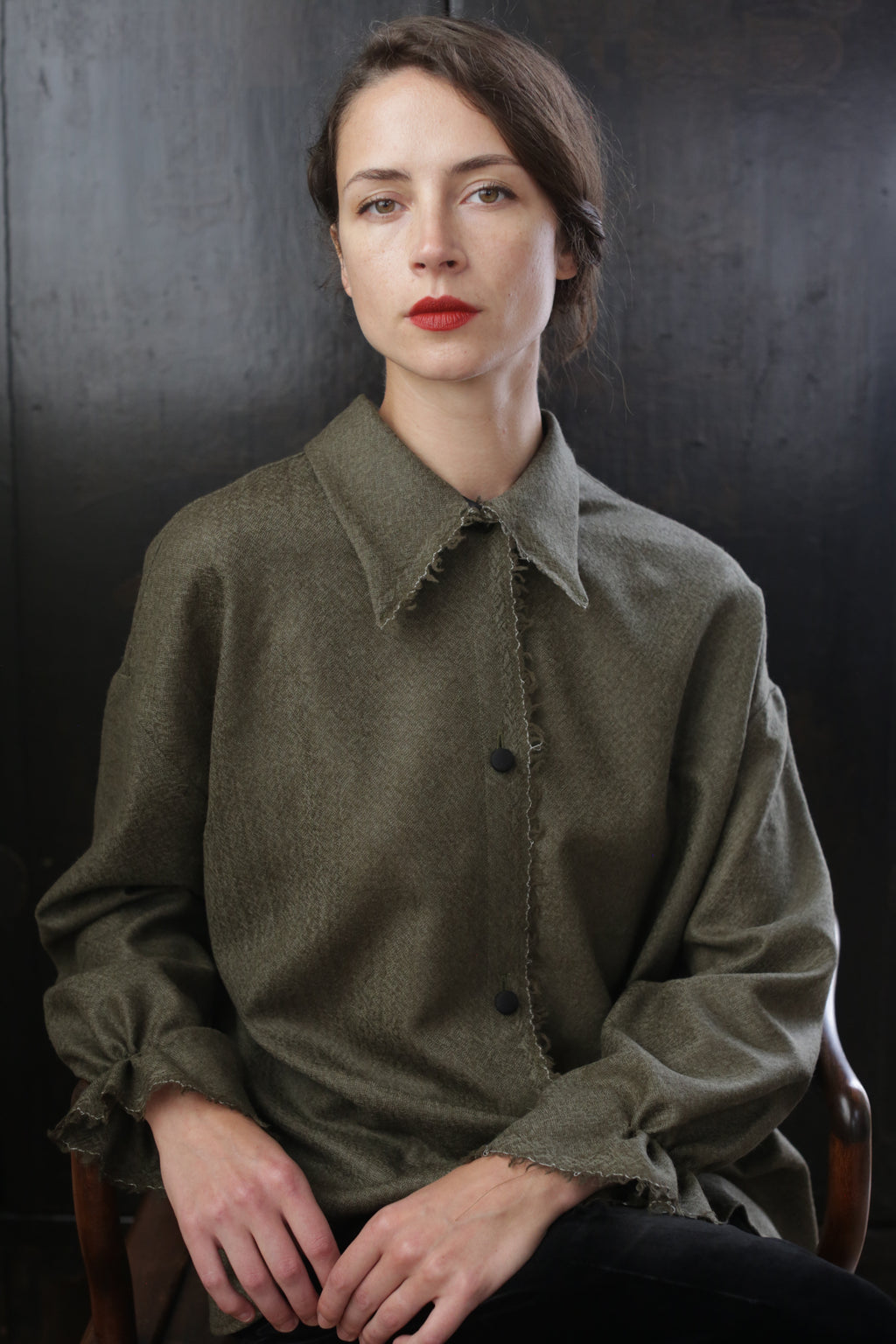 A woman with long, dark hair and red lipstick sits on a wooden chair against a dark background. She wears an oversized, olive-green Painter's Shirt - Japanese Wool Gauze | Olive with frayed edges and holds her hands in her lap, gazing directly at the camera with a neutral expression.