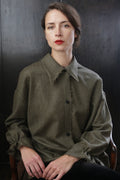 A woman with long, dark hair and red lipstick sits on a wooden chair against a dark background. She wears an oversized, olive-green Painter's Shirt - Japanese Wool Gauze | Olive with frayed edges and holds her hands in her lap, gazing directly at the camera with a neutral expression.