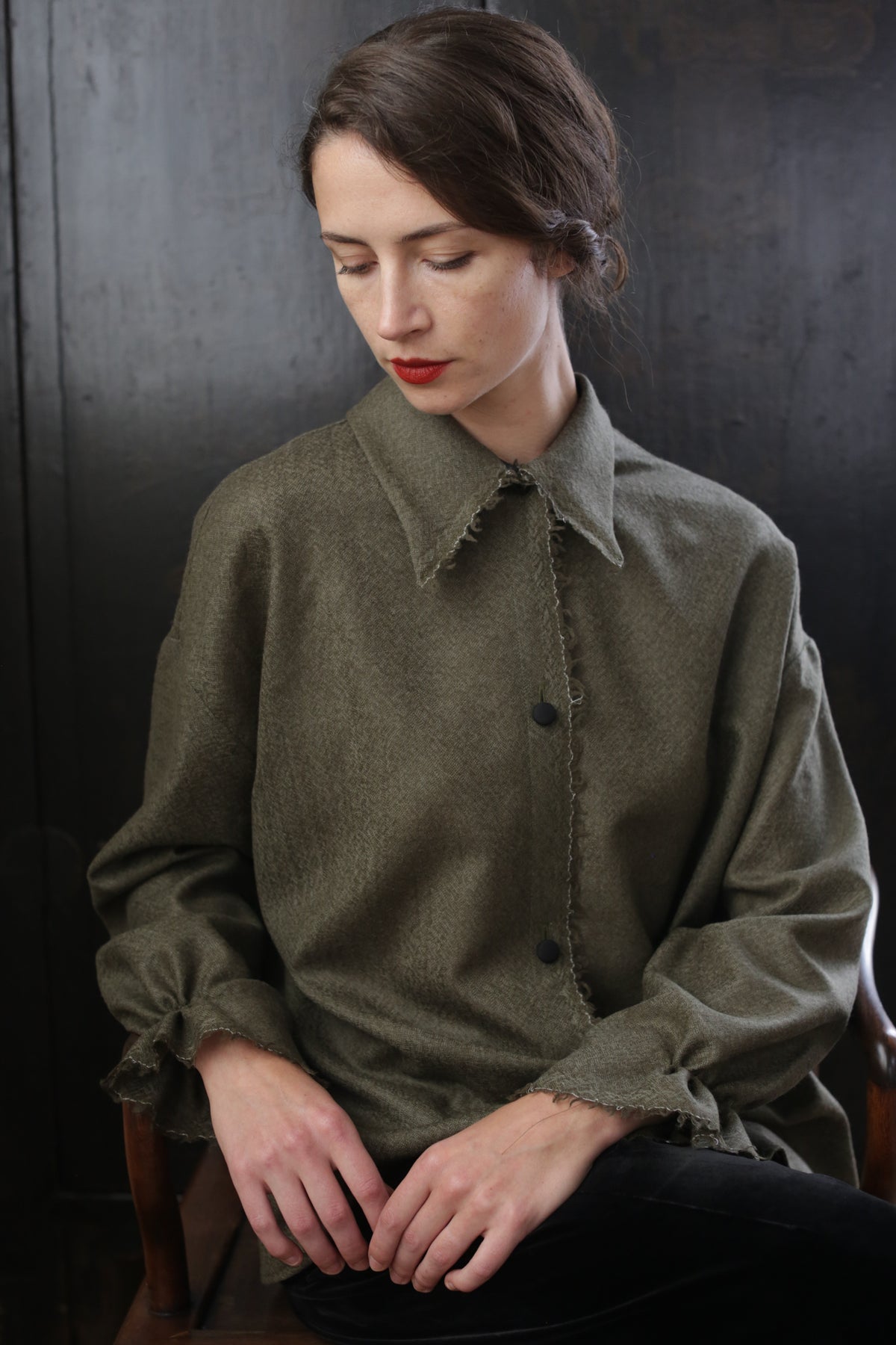 A person with dark brown hair, wearing red lipstick, and dressed in a loose-fitting, greenish-brown Painter's Shirt - Japanese Wool Gauze | Olive is sitting in a dark room with their head slightly tilted downward and eyes closed, creating a pensive or contemplative expression.
