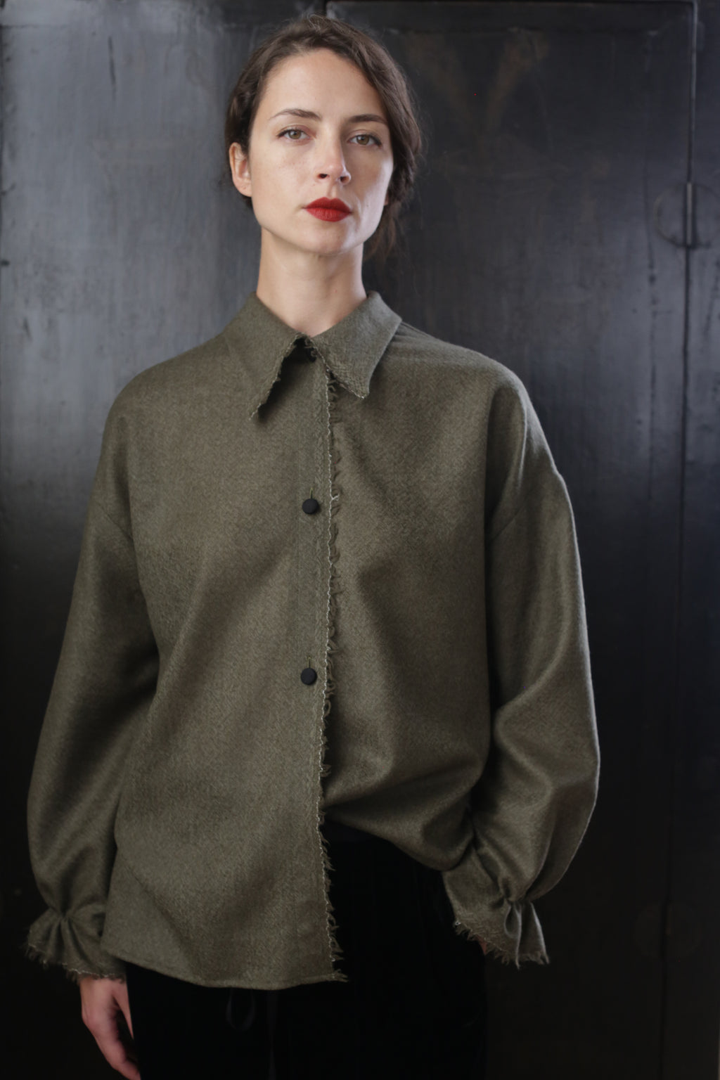 A person with short dark hair and red lipstick stands against a dark background, wearing a loose-fitting Painter's Shirt - Japanese Wool Gauze | Olive with frayed edges and ruffled cuffs. The expression is neutral or slightly serious.