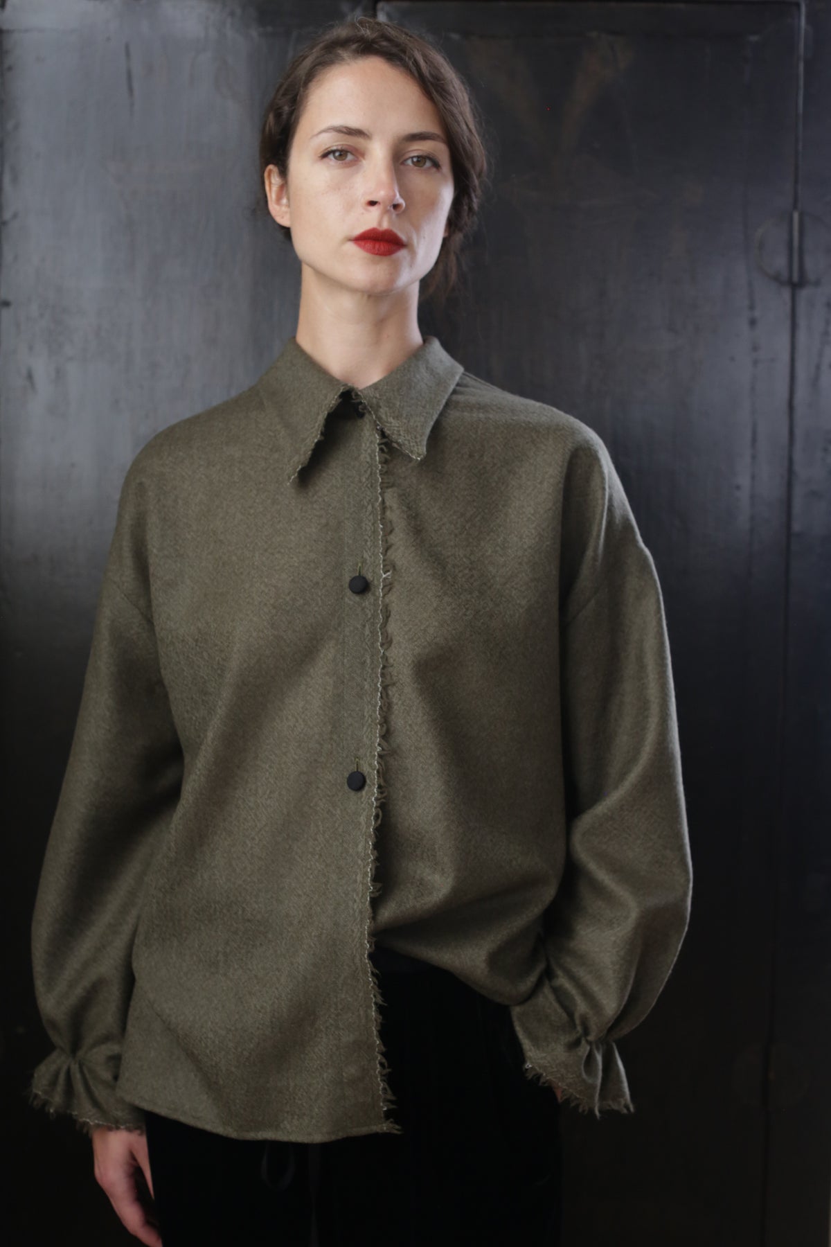 A person with short dark hair and red lipstick stands against a dark background, wearing a loose-fitting Painter's Shirt - Japanese Wool Gauze | Olive with frayed edges and ruffled cuffs. The expression is neutral or slightly serious.