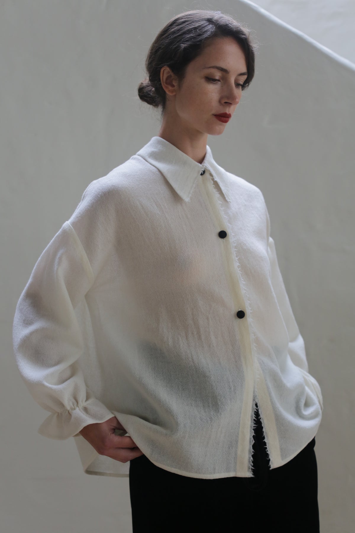 A person with dark hair is standing against a light gray background. They are wearing a Painter's Shirt - Japanese Wool Gauze | Undyed with black buttons and black pants. They have a neutral expression and their hands placed casually on their hips.