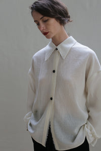 A person stands against a plain background, wearing an oversized Painter's Shirt - Japanese Wool Gauze | Undyed, featuring black buttons and frayed edges. Their short, dark hair is styled neatly, and they have their hands in their pockets while gazing downwards with a neutral expression.