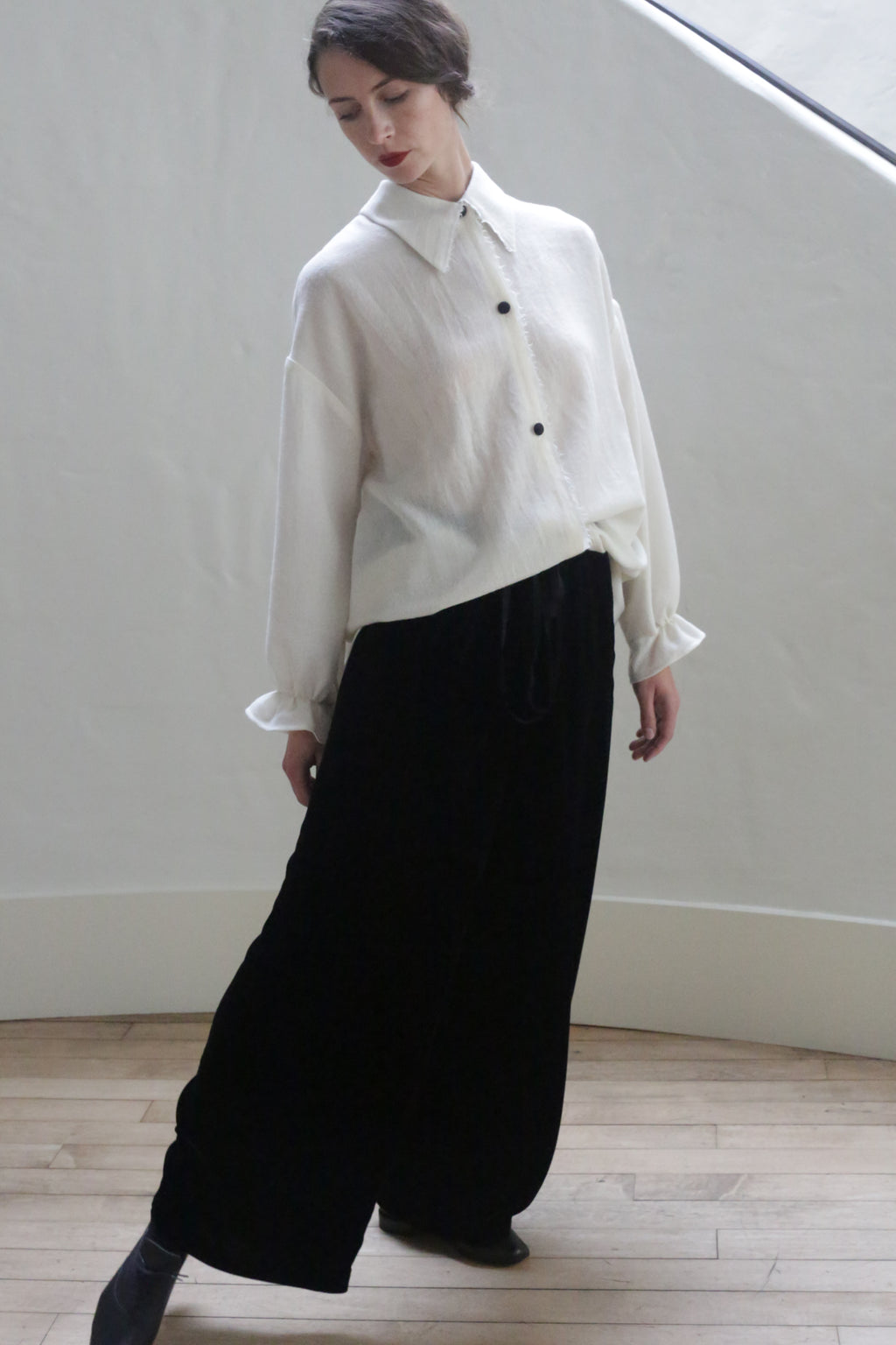 A person stands indoors on a wooden floor against a white wall, wearing the Painter's Shirt - Japanese Wool Gauze | Undyed and wide black pants made from Japanese wool gauze. They look down and slightly to the side with a relaxed posture, their hair tied back, showcasing elegant handmade clothing with sustainable design.
