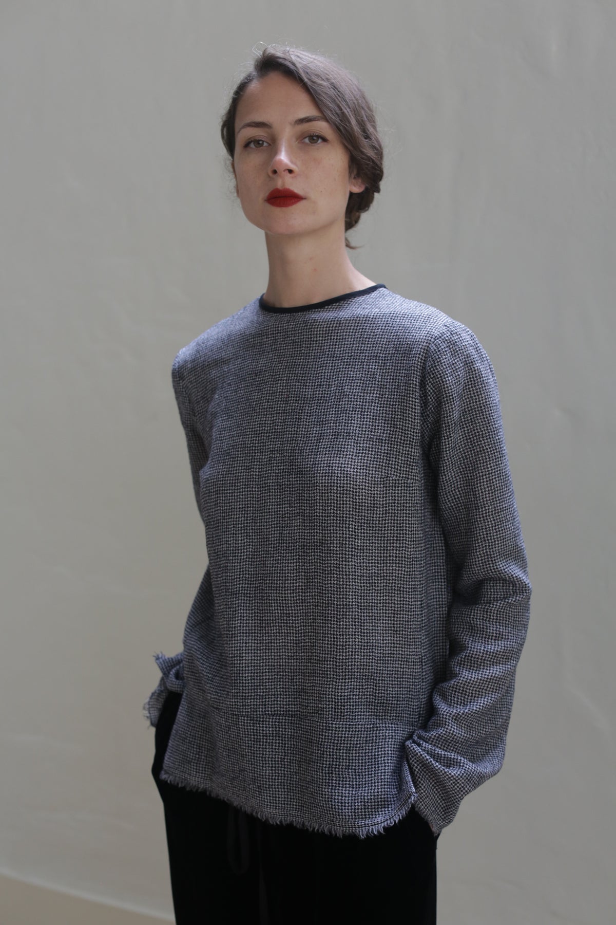 A person with short brown hair, wearing a Crewneck shirt - Japanese Wool Gauze | BW Houndstooth in gray, stands against a plain, light-colored background. The individual gazes at the camera with a neutral expression, hands partially in the pockets of dark pants. The shirt's Japanese wool gauze adds a touch of sustainable design to the overall look.