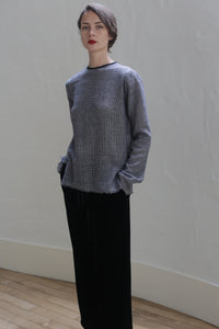 A person with short, dark hair is standing against a plain white wall. They are wearing a gray Crewneck shirt - Japanese Wool Gauze | BW Houndstooth and black pants. Their hands are tucked into their pockets, and they are looking forward with a neutral expression. Light wooden flooring is visible.