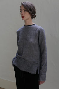 A person stands against a light-colored wall, looking to the side. They have short, dark hair styled in a bun and wear a Crewneck shirt - Japanese Wool Gauze | BW Houndstooth paired with black pants. Their outfit includes a gray textured sweater made from Japanese wool gauze. They have a calm and contemplative expression.