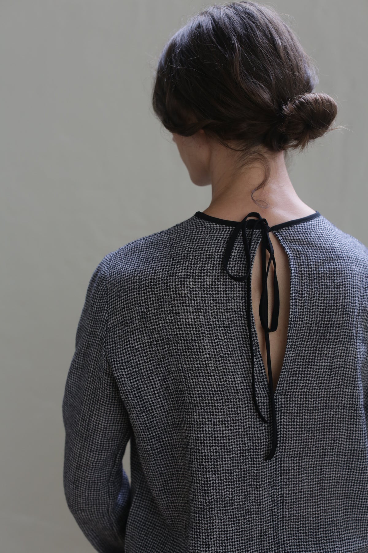 A woman with brown hair styled in a low bun is seen from behind. She is wearing the Crewneck shirt - Japanese Wool Gauze | BW Houndstooth, which has an open back tied with black strings. The background is plain and neutral, highlighting the sustainable design of her outfit.