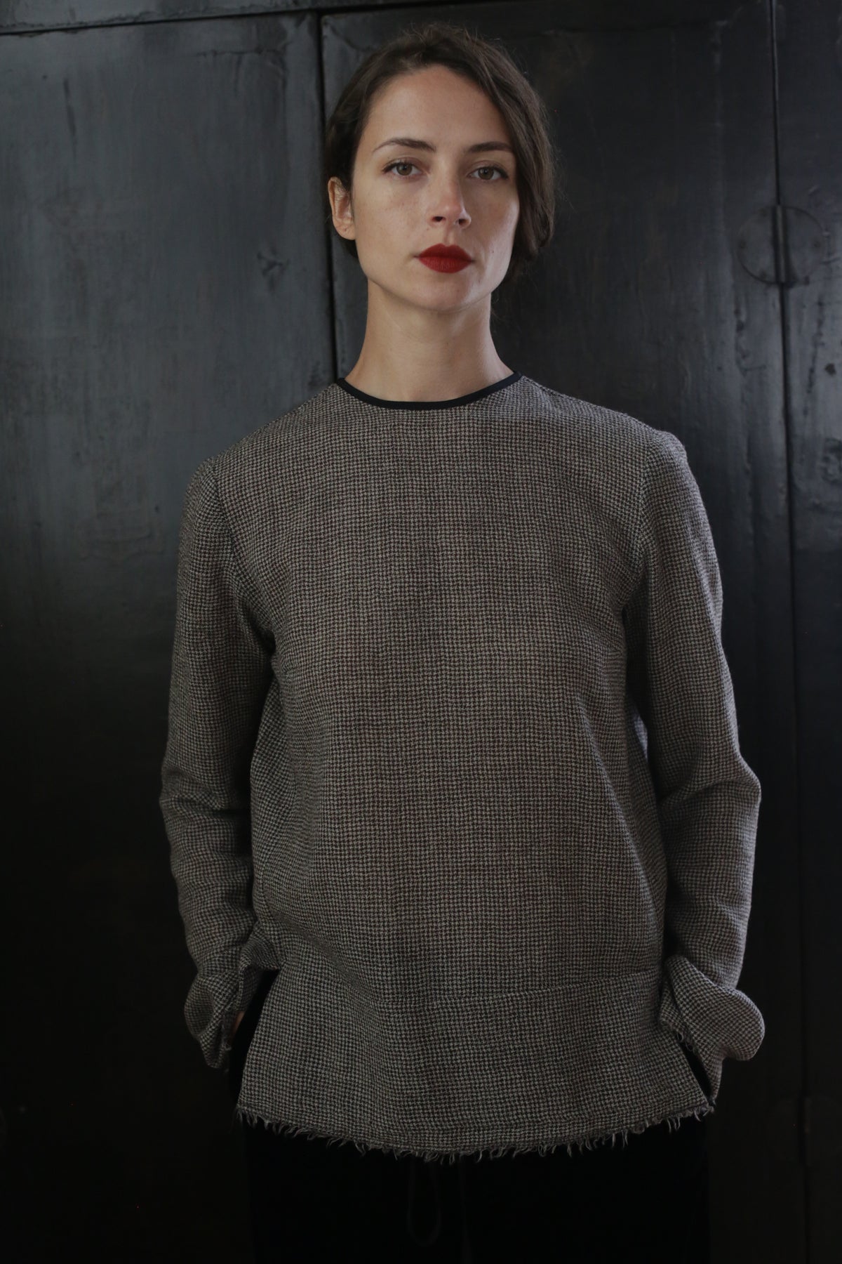A woman with shoulder-length brown hair stands against a dark, textured background. Wearing a Crewneck shirt - Japanese Wool Gauze | Brown Houndstooth, she has her hands in her pockets. She gazes at the camera with a neutral expression and wears bright red lipstick, embodying sustainable design.