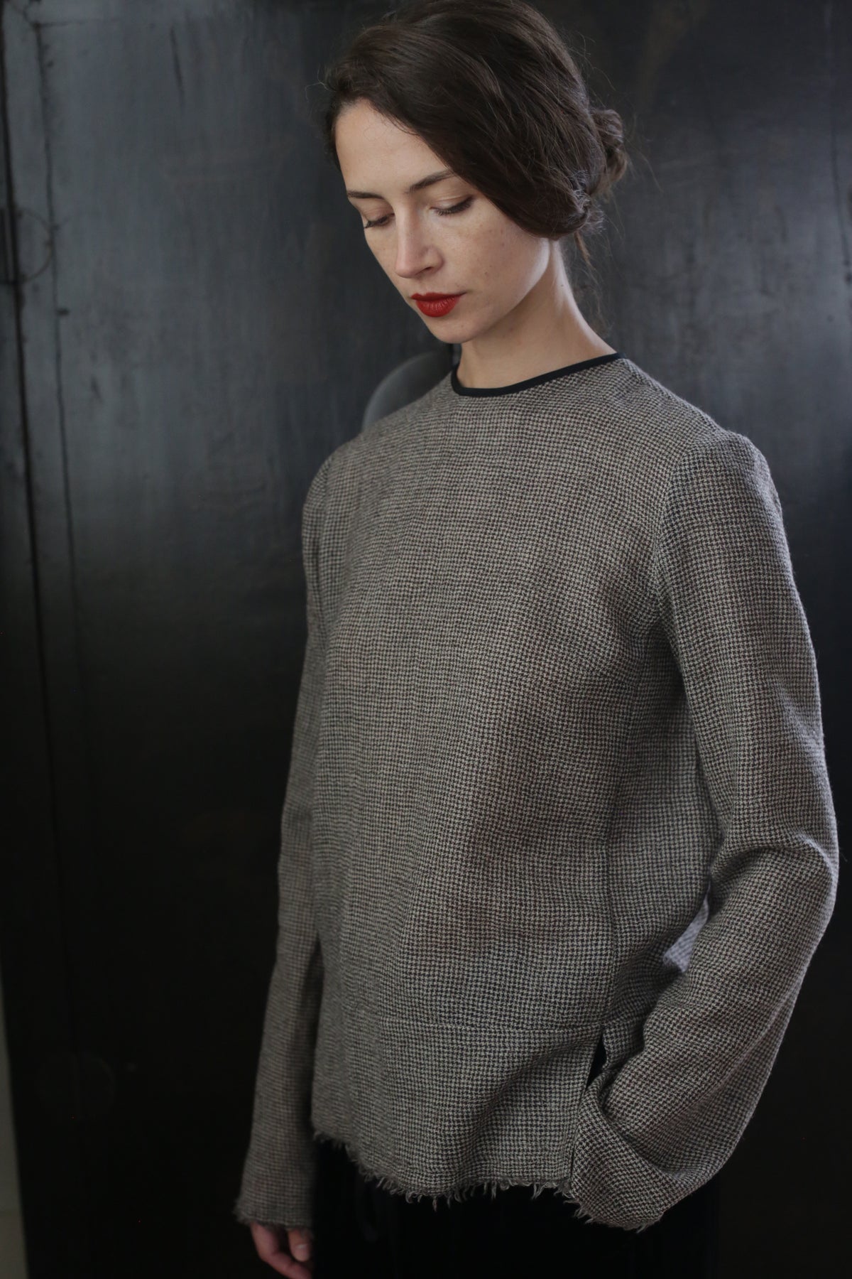 A person with long hair wearing a handmade, neutral-toned, Crewneck shirt - Japanese Wool Gauze | Brown Houndstooth stands against a dark background. They are looking downward with their head slightly tilted, and one hand is placed in their pocket. Their expression appears contemplative.