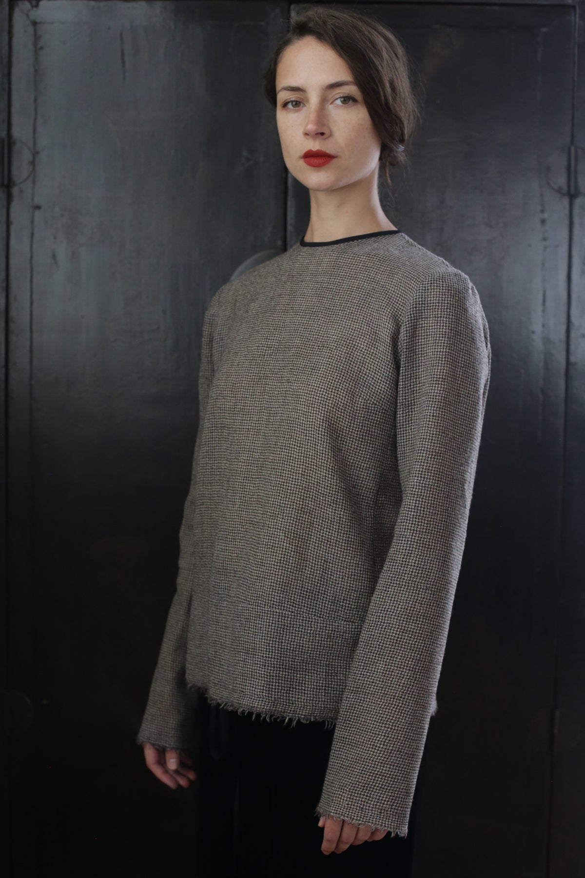 A person stands against a dark background wearing a textured, long-sleeved Crewneck shirt - Japanese Wool Gauze | Brown Houndstooth. They have medium-length hair styled neatly and wear red lipstick. Their expression is neutral, and they face directly towards the camera, showcasing a touch of sustainable design.