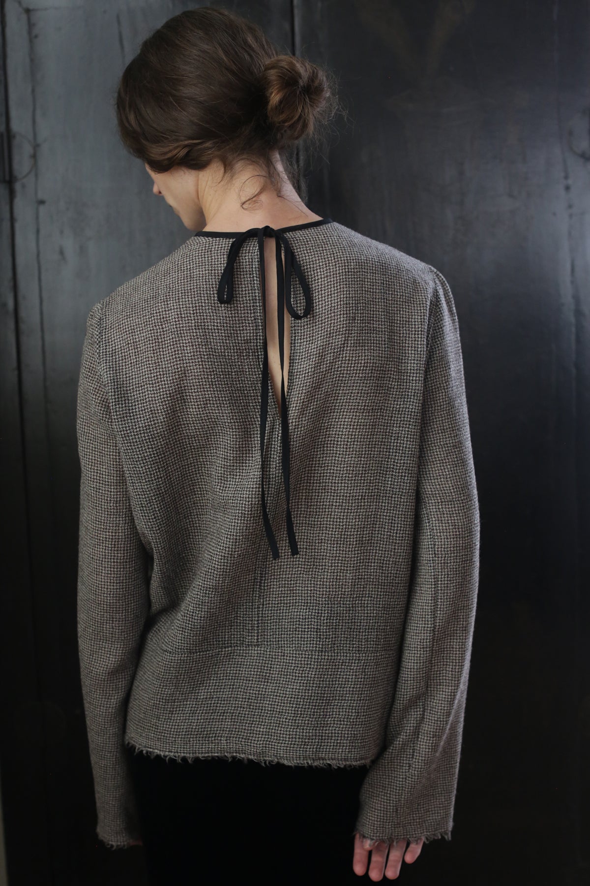A person with brown hair styled in a loose bun is facing away from the camera. They are wearing a Crewneck shirt - Japanese Wool Gauze | Brown Houndstooth featuring a black ribbon tie at the back, made from sustainable design principles. The background is dark and minimalist, accentuating the focus on the handmade clothing and hairstyle.