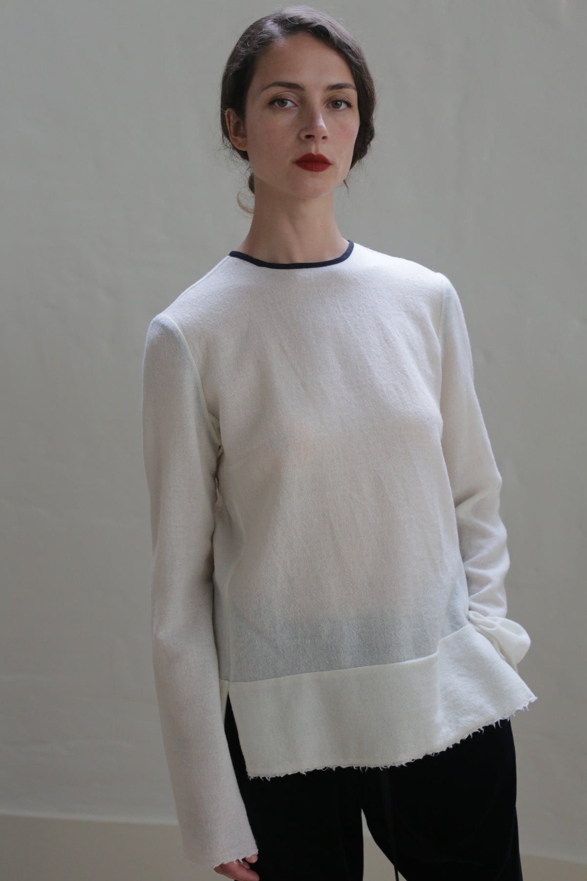 A woman with dark hair tied back is standing against a plain background. She is wearing a long-sleeved crewneck shirt - Japanese Wool Gauze | Undyed with dark trim and black pants. Her arms are at her sides, and she is looking directly at the camera with a neutral expression, embodying sustainable design.