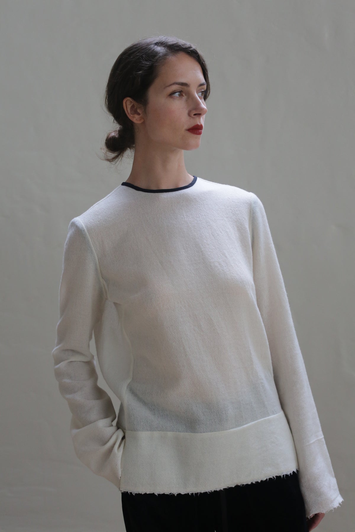 A woman with dark hair is styled in a low bun, looking off to the side with a soft, pensive expression. She is wearing a Crewneck shirt - Japanese Wool Gauze | Undyed with black accents on the cuffs. The background is a plain, neutral color, keeping the focus on her.