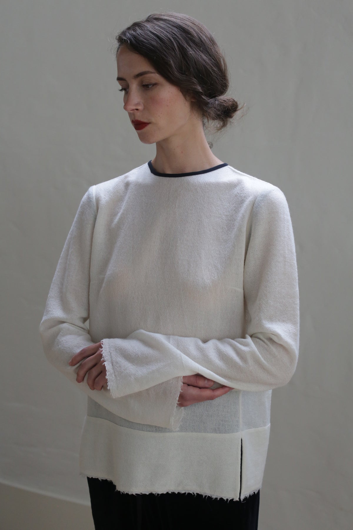 A person with long dark hair and red lipstick stands against a plain background. They are wearing a loose-fitting, long-sleeved **Crewneck shirt - Japanese Wool Gauze | Undyed** and dark pants. Their head is slightly tilted to the side, and their arms are crossed in a relaxed manner, embodying sustainable design.
