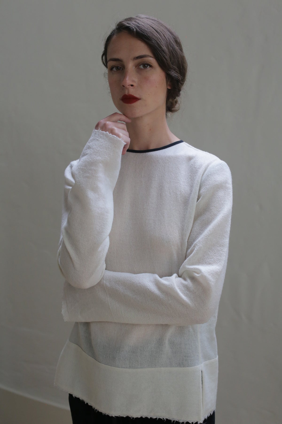 A woman with dark hair styled in an updo is standing against a plain background. She is wearing a Crewneck shirt - Japanese Wool Gauze | Undyed, reflecting sustainable design. Her left arm is crossed, with her right hand touching her chin as she looks straight at the camera with a serious expression.