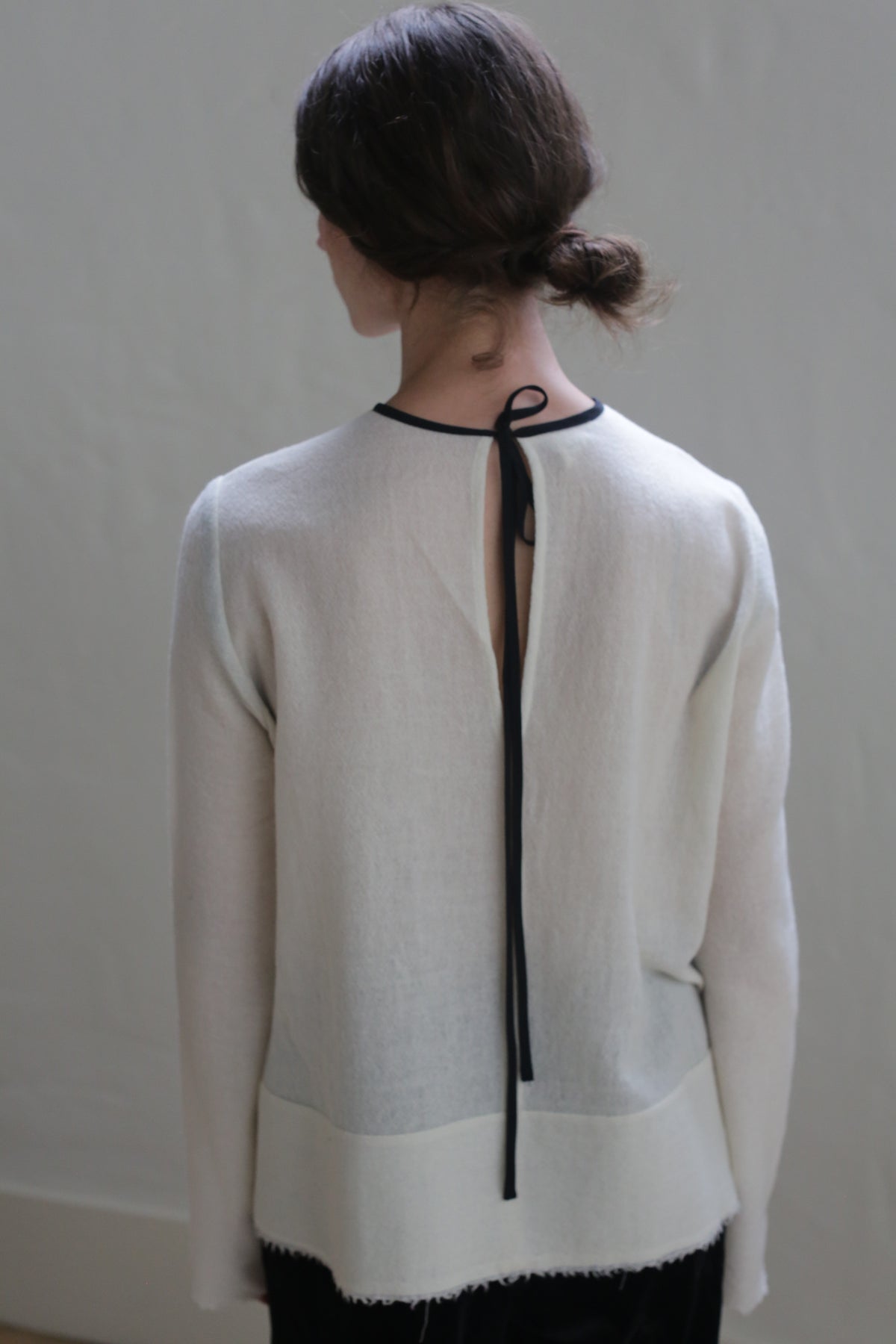 A person with dark hair tied in a low bun is wearing a Crewneck shirt - Japanese Wool Gauze | Undyed, featuring a back opening tied with a black ribbon. They are facing away from the camera against a neutral background.