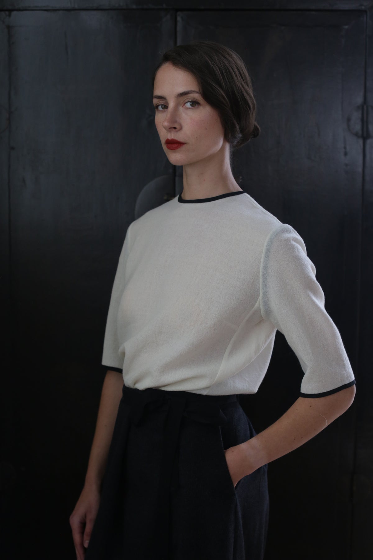 A woman with short dark hair stands against a dark background. She is wearing the Crewneck Tee Japanese Wool Gauze - Undyed and a dark skirt made from Japanese wool gauze, with one hand in a pocket. Her expression is neutral, and she looks directly at the camera.