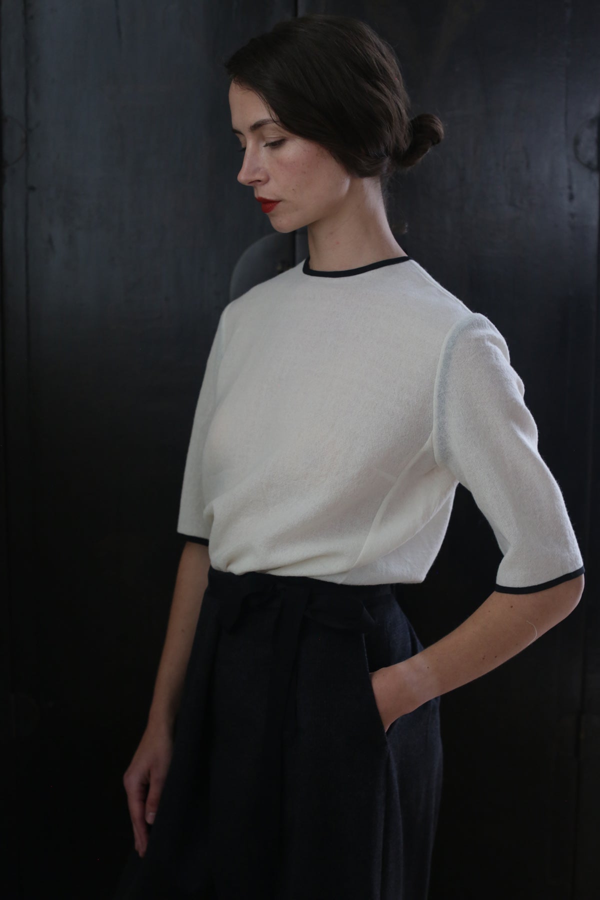 A woman with dark hair pulled back in a bun stands against a dark background. She is wearing a white, handmade Crewneck Tee Japanese Wool Gauze - Undyed with black trim around the neckline and sleeves, and high-waisted black pants. Her eyes are closed, and her expression is calm and thoughtful, embodying sustainable design principles.