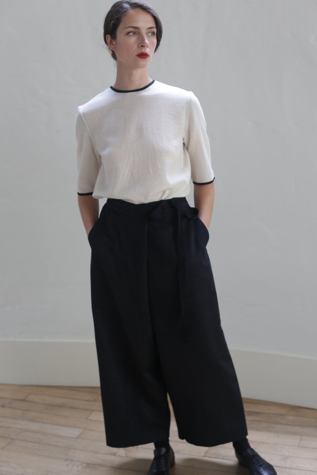 A person with short dark hair stands against a plain, light grey wall. They are wearing a Crewneck Tee Japanese Wool Gauze - Undyed and loose black wide-leg trousers made from Japanese wool gauze, with hands in pockets. The image is taken indoors on a wooden floor, showcasing sustainable design.
