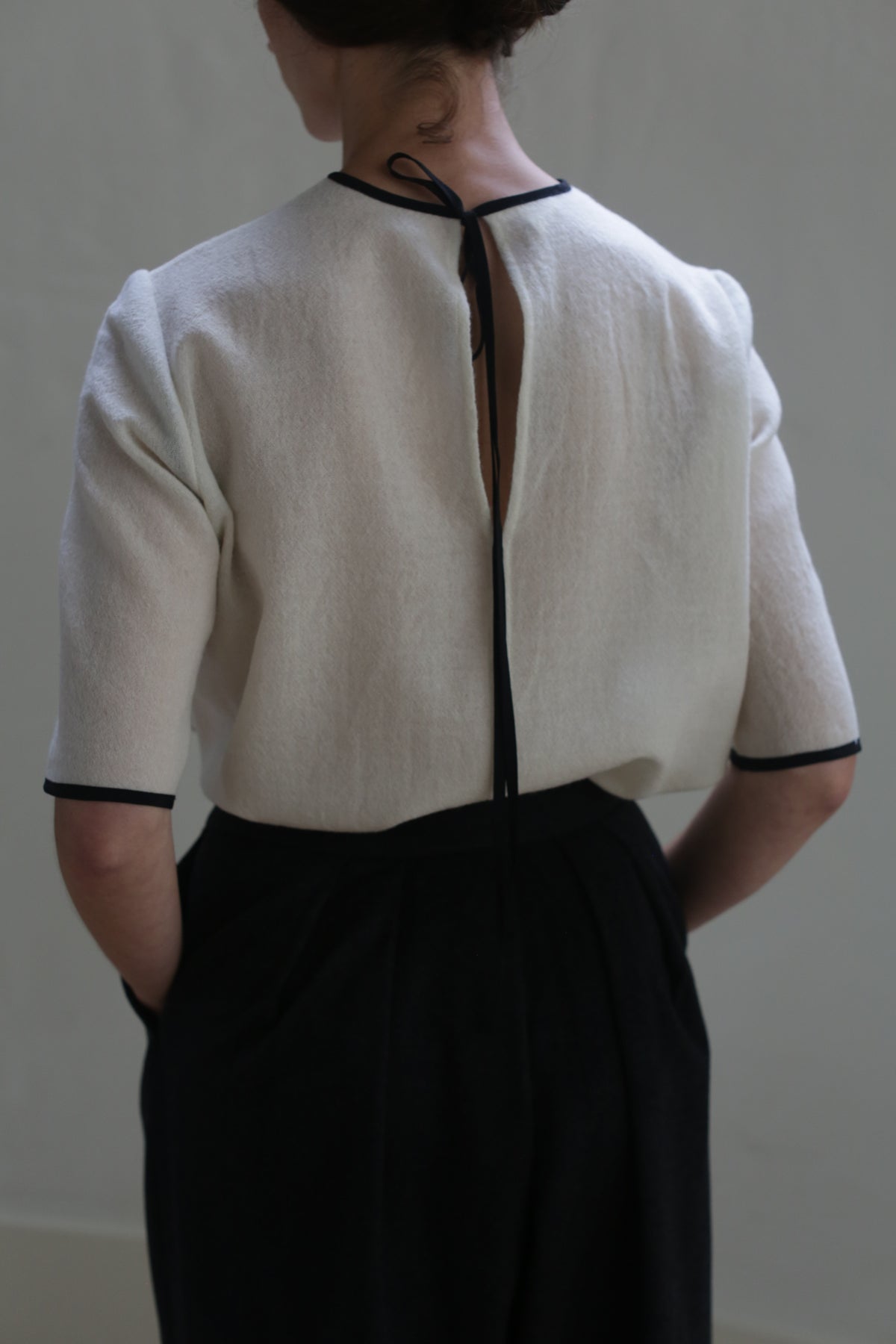 Person standing with their back facing the camera, wearing a Crewneck Tee Japanese Wool Gauze - Undyed, with black trim and a long black tie closure at the back of the neck. They also have their hands in the pockets of high-waisted black pants made from Japanese wool gauze. The background is light gray and plain, highlighting sustainable design.
