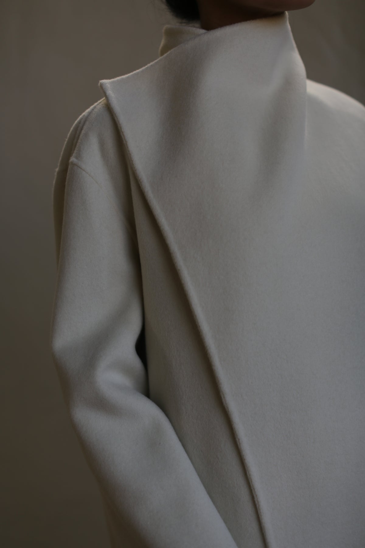 A close-up of a person wearing a sophisticated, light gray Winston Long Double-face. The coat features a unique, asymmetrical design and smooth, refined fabric with a relaxed construction. The background is softly blurred, emphasizing the coat's texture and design details.