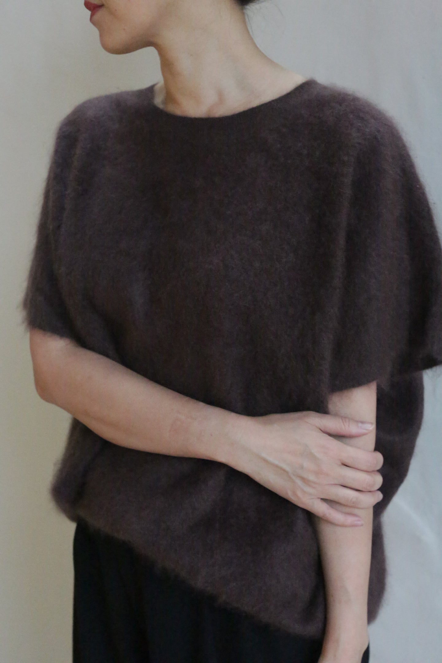A person wearing the Salento | Mink loose, short-sleeved brushed cashmere sweater holds their left arm with their right hand and faces slightly away from the camera against a plain background.