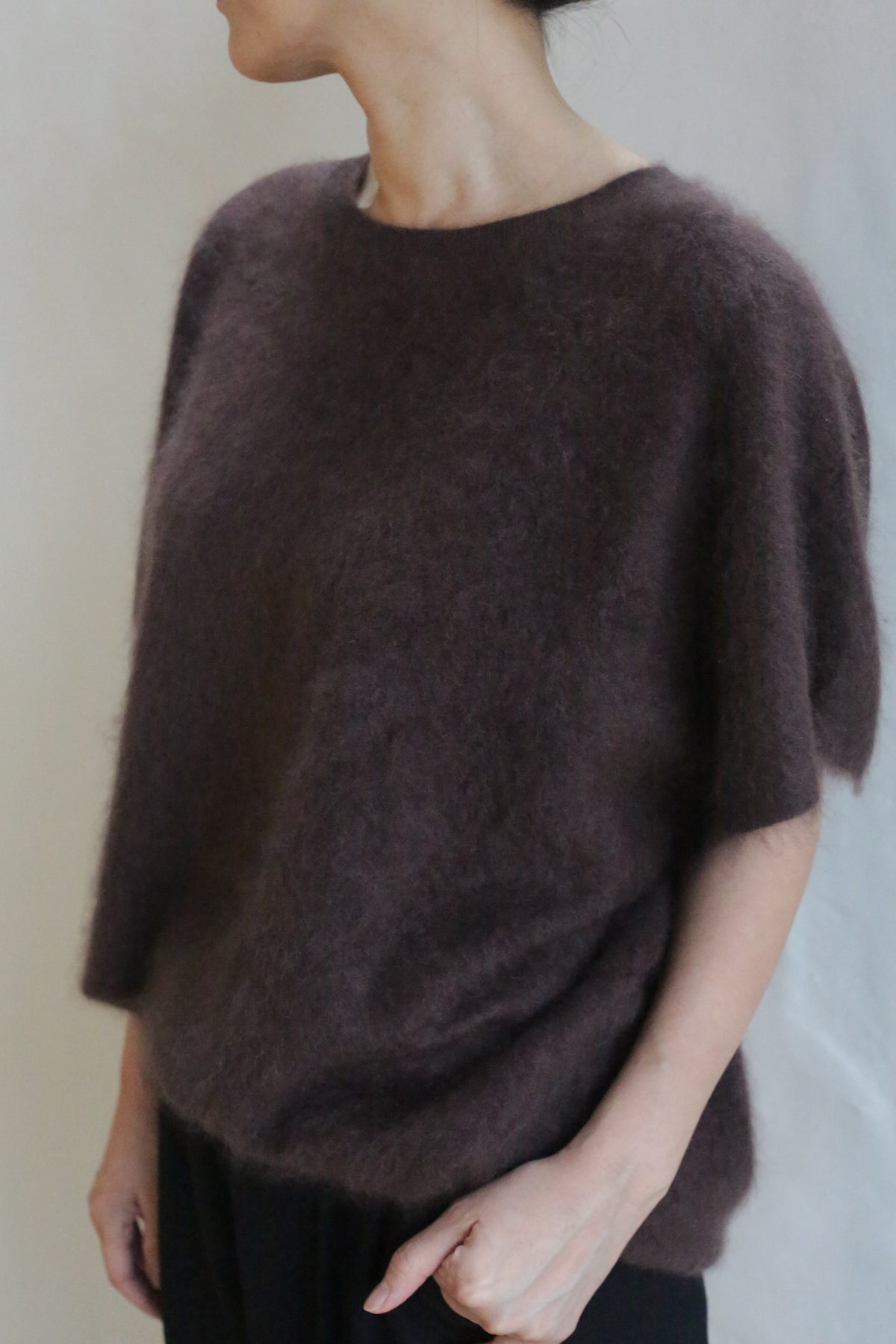 A person wears the Salento | Mink loose, short-sleeved, dark brown brushed cashmere sweater with black pants, standing against a plain light background and facing slightly away from the camera with one hand in their pocket.