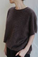 Load image into Gallery viewer, A person wears the Salento | Mink loose, short-sleeved, dark brown brushed cashmere sweater with black pants, standing against a plain light background and facing slightly away from the camera with one hand in their pocket.