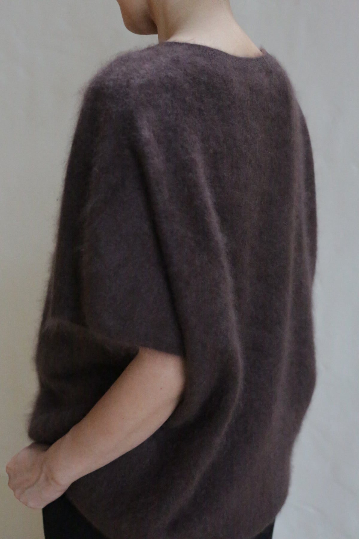 A person stands with their back and side visible, wearing the Salento | Mink—a loose, fuzzy brushed cashmere short-sleeved sweater—against a plain light background.
