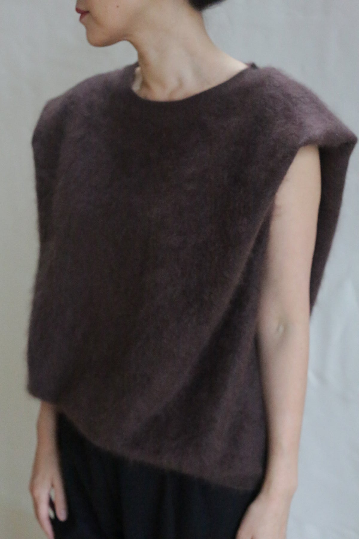 Wearing the Salento | Mink—a sleeveless, loose-fit brushed cashmere dark brown top—paired with black pants, a person stands against a light background, looking to the side with their face partially out of frame.
