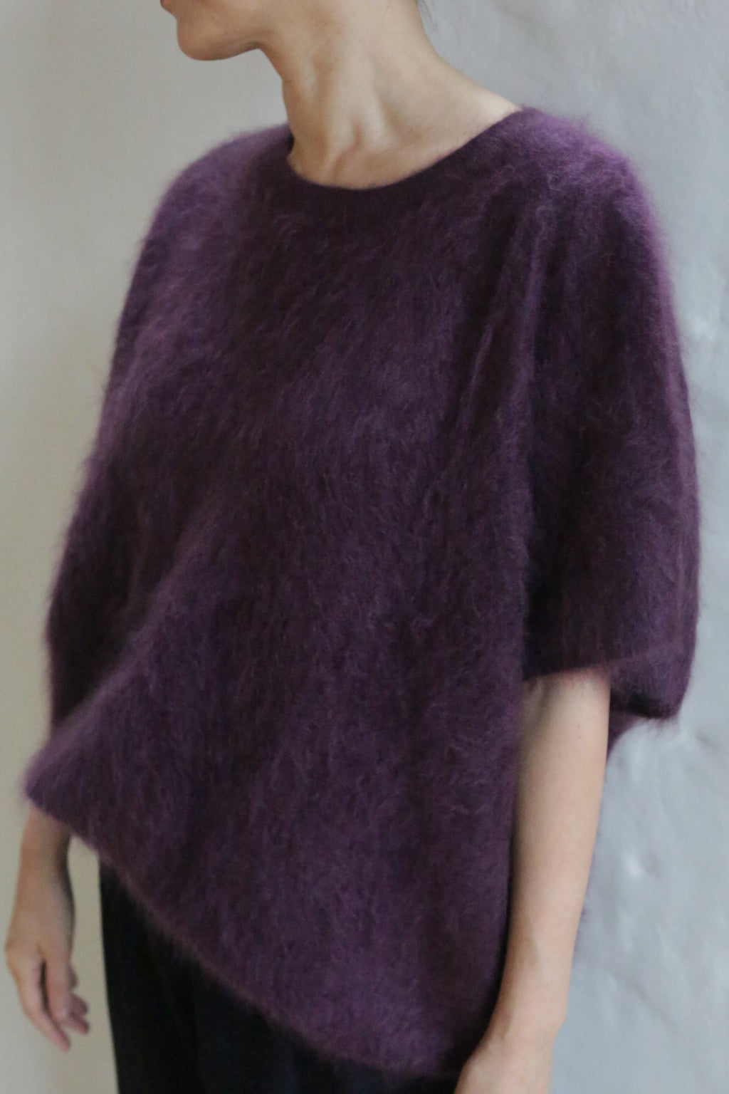 Wearing the Salento | Aubergine loose-fitting, short-sleeved brushed cashmere sweater and black pants, a person stands against a light wall with their head slightly turned out of frame.
