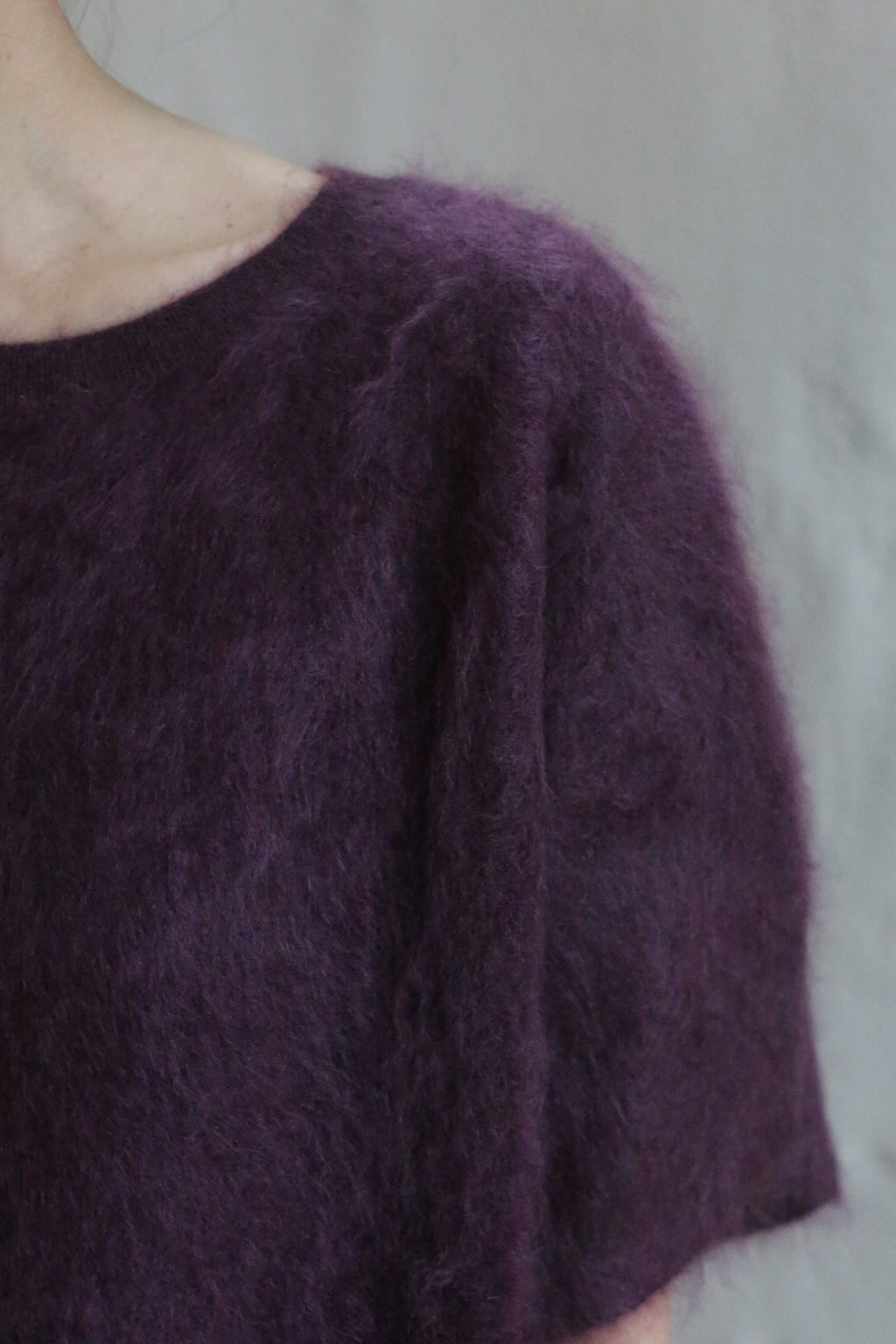 Close-up of a person wearing the Salento | Aubergine short-sleeved, brushed cashmere sweater. Their neck and part of their left shoulder are visible against a neutral background.