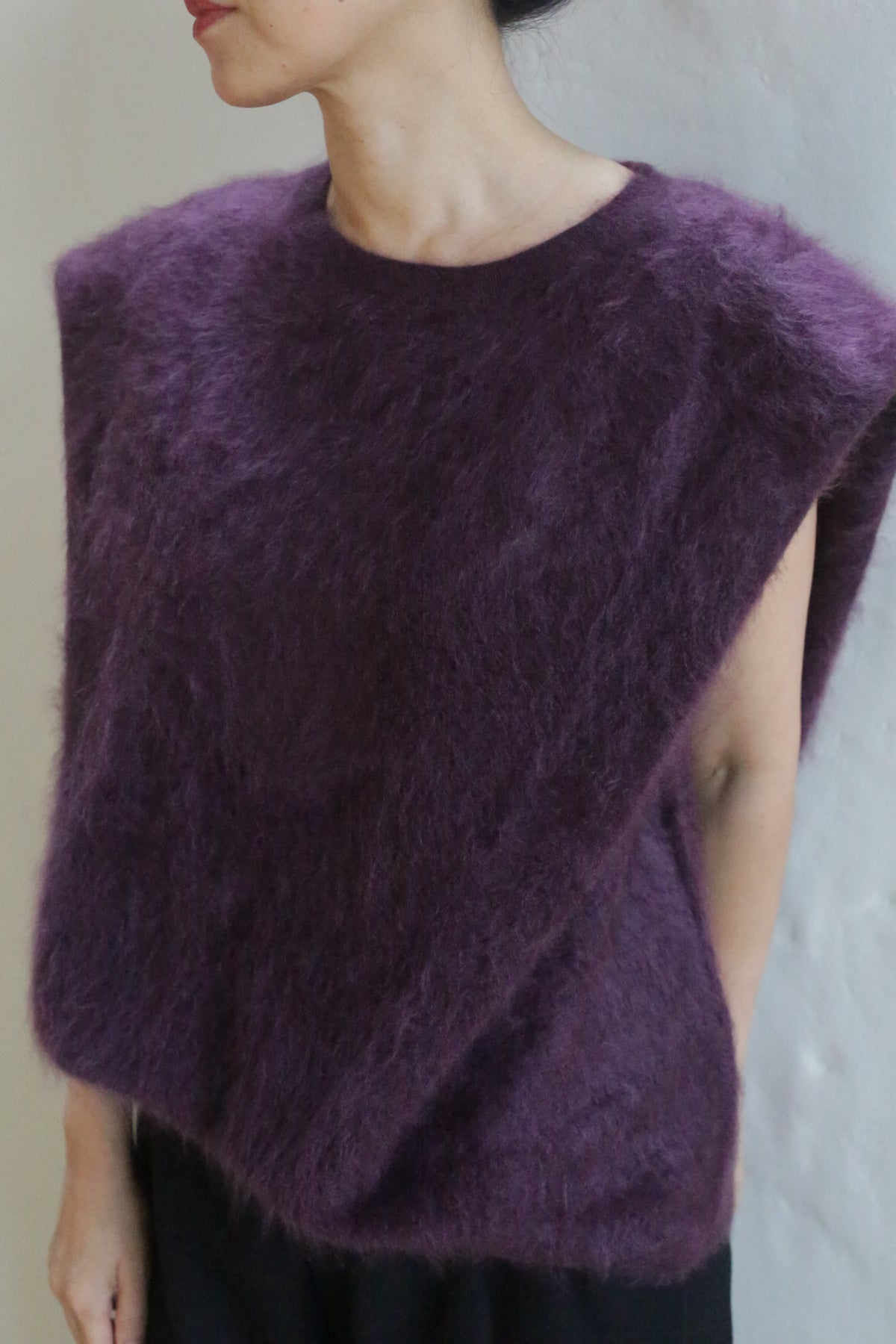 A person stands against a plain light background wearing the Salento | Aubergine sleeveless brushed cashmere top with a wide neckline, head turned to the side and hands behind their back.