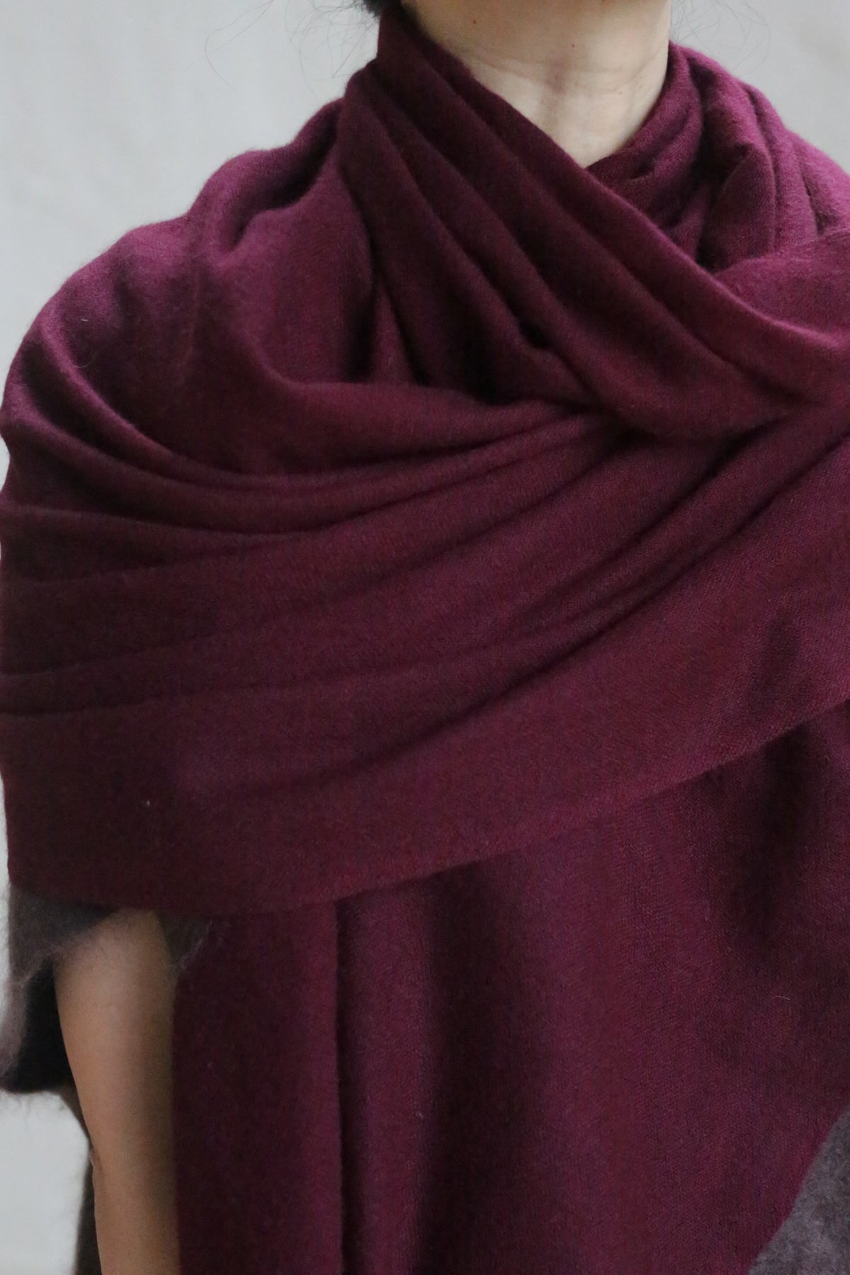 A person drapes the Tissue Weight Cashmere Scarf in Garnet, sized 36" x 82", around their shoulders with layered folds, showing only their lower face, neckline, and upper torso.